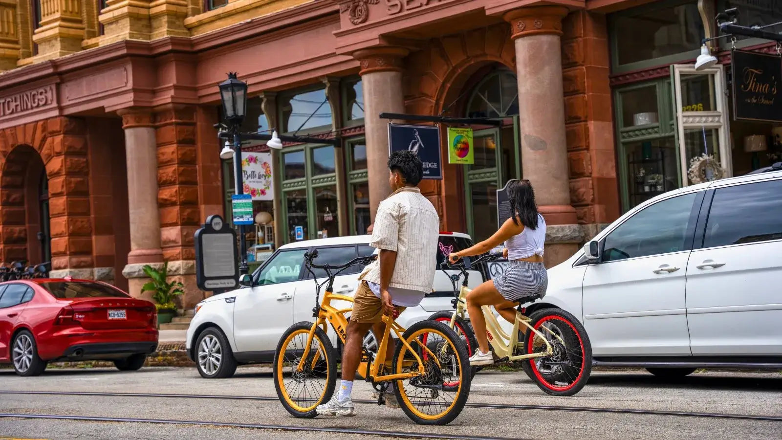 Two people riding colorful electric beach cruiser bikes past historic buildings in a city street
