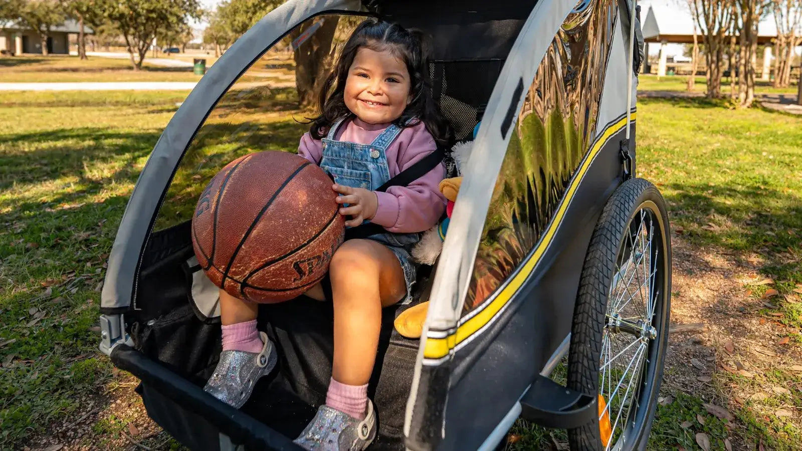 Smiling child sitting inside a bike trailer holding a basketball, demonstrating comfort and safety for young passengers.