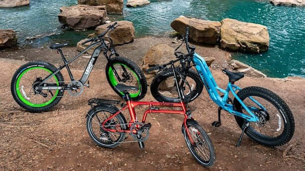 Red and blue electric bikes parked by a scenic lakeside with rocks and clear water