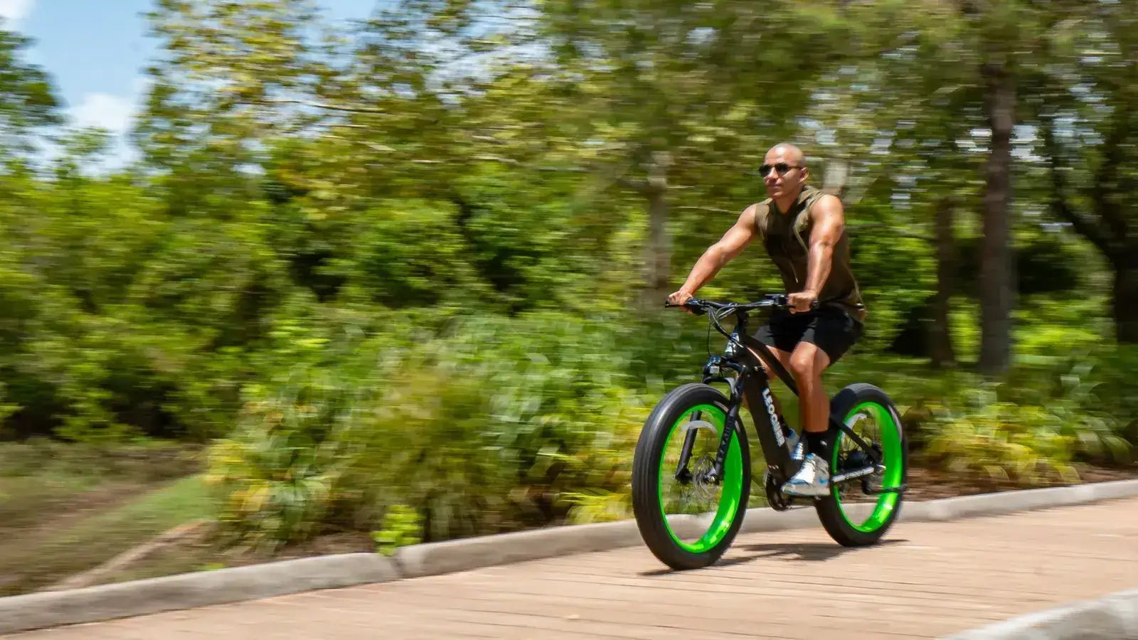 Man riding a fat tire electric bike at high speed on a paved trail surrounded by greenery