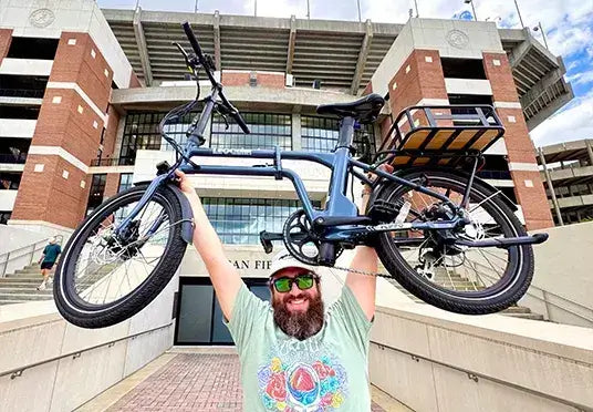 Man holding an electric bike overhead in front of a stadium, celebrating California ebike incentive success