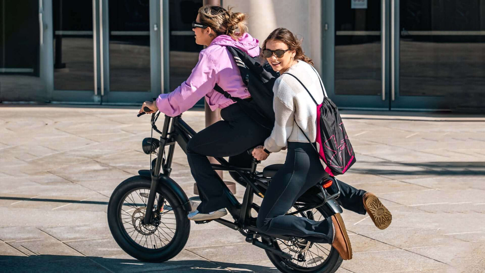 Two women riding together on a compact electric bike in an urban area, sharing the seat while smiling.