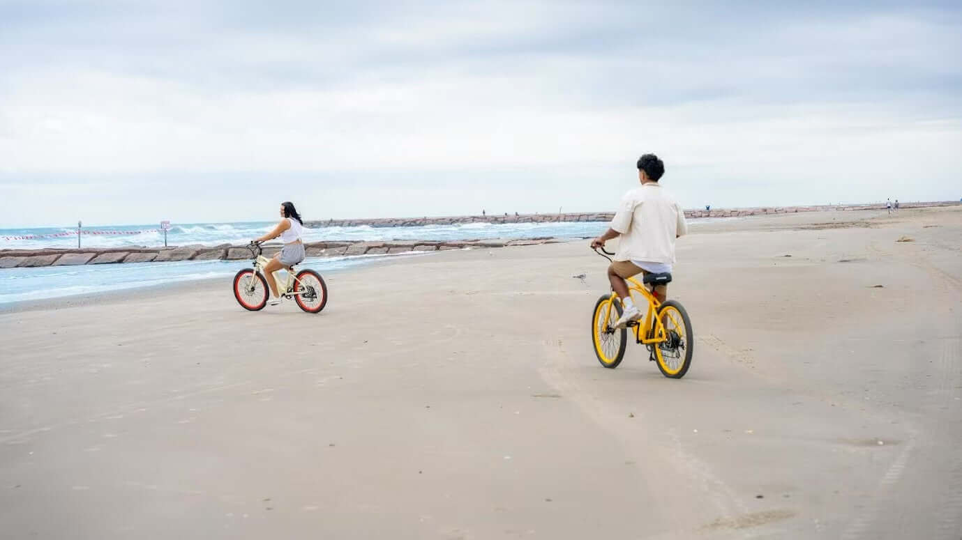 Two riders on cruiser e-bikes riding along a beach path in 2025