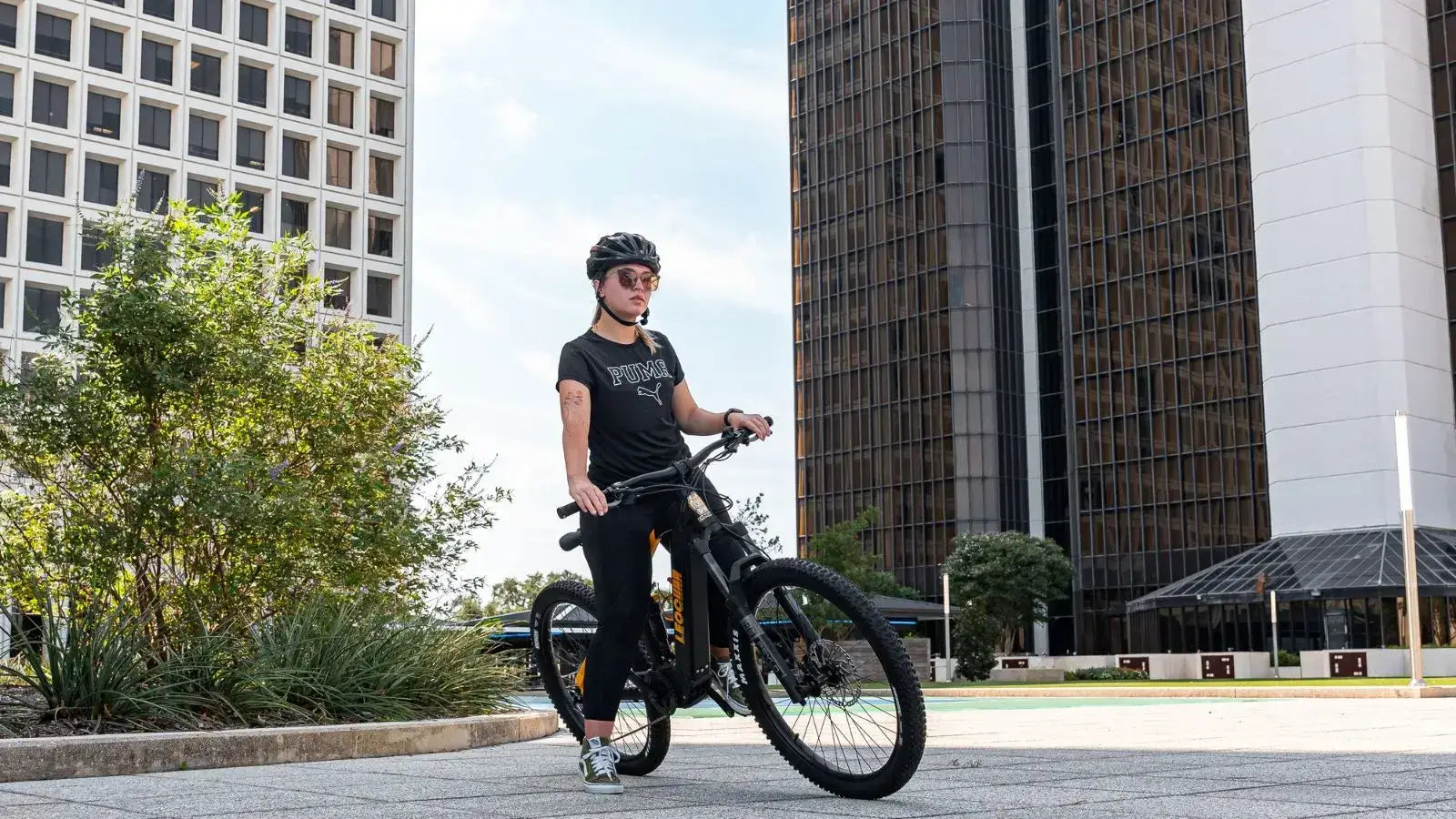 Rider on a black electric dirt bike with pedals, cruising in an urban setting with tall modern buildings in the background