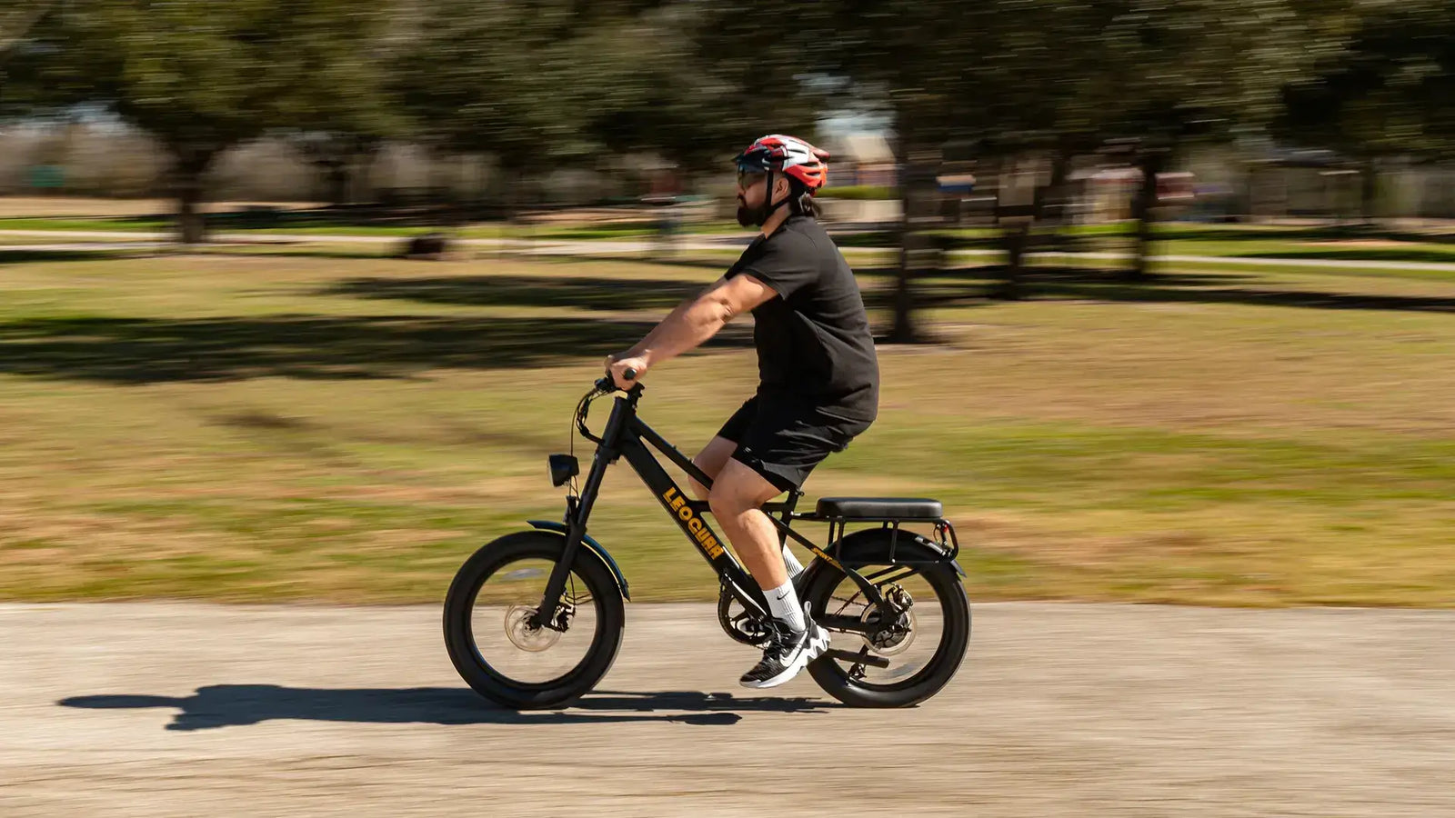 Person riding a compact 20-inch electric bike through a park on a sunny day, demonstrating portability and ease of use