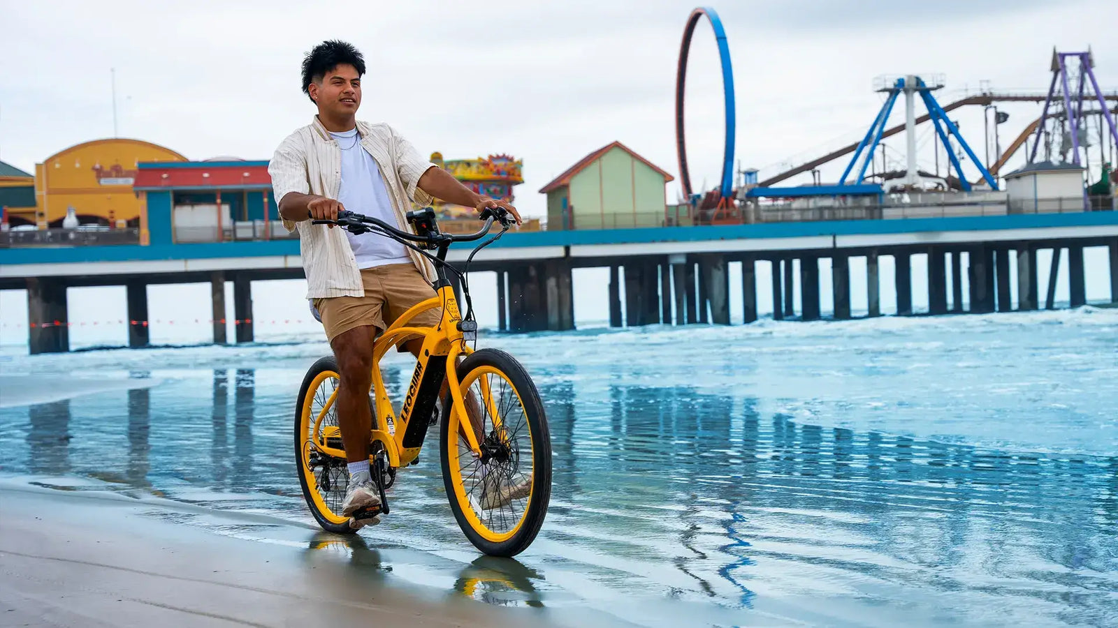 Man riding a bright yellow men's beach cruiser e-bike along a seaside boardwalk with colorful buildings and a pier in the background.