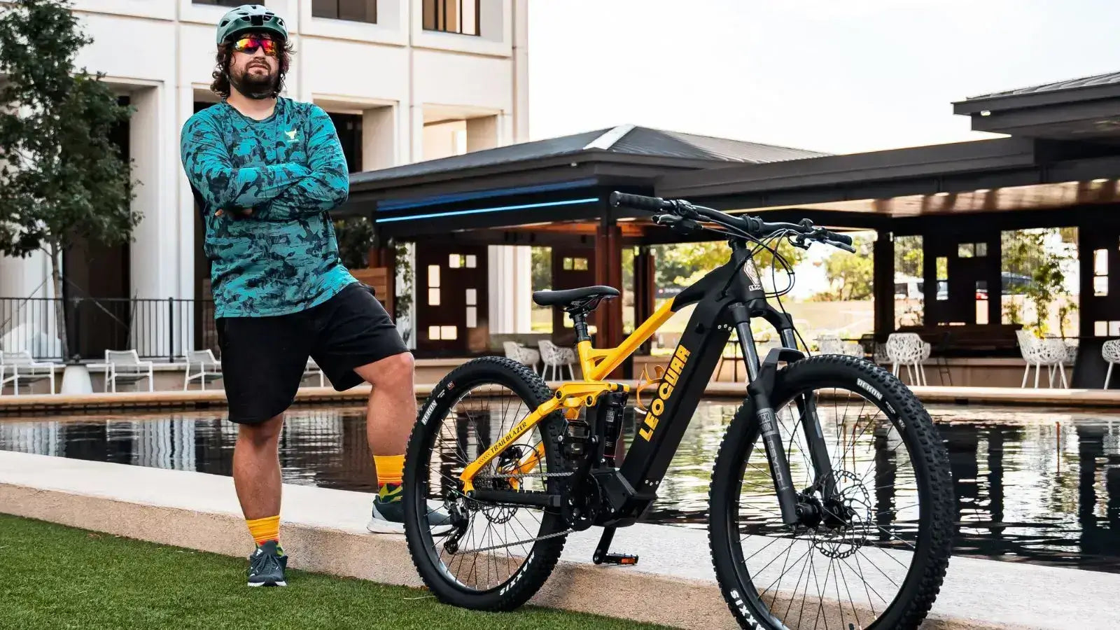 Man standing next to a yellow all-terrain electric mountain bike in front of a modern building with a reflective pool