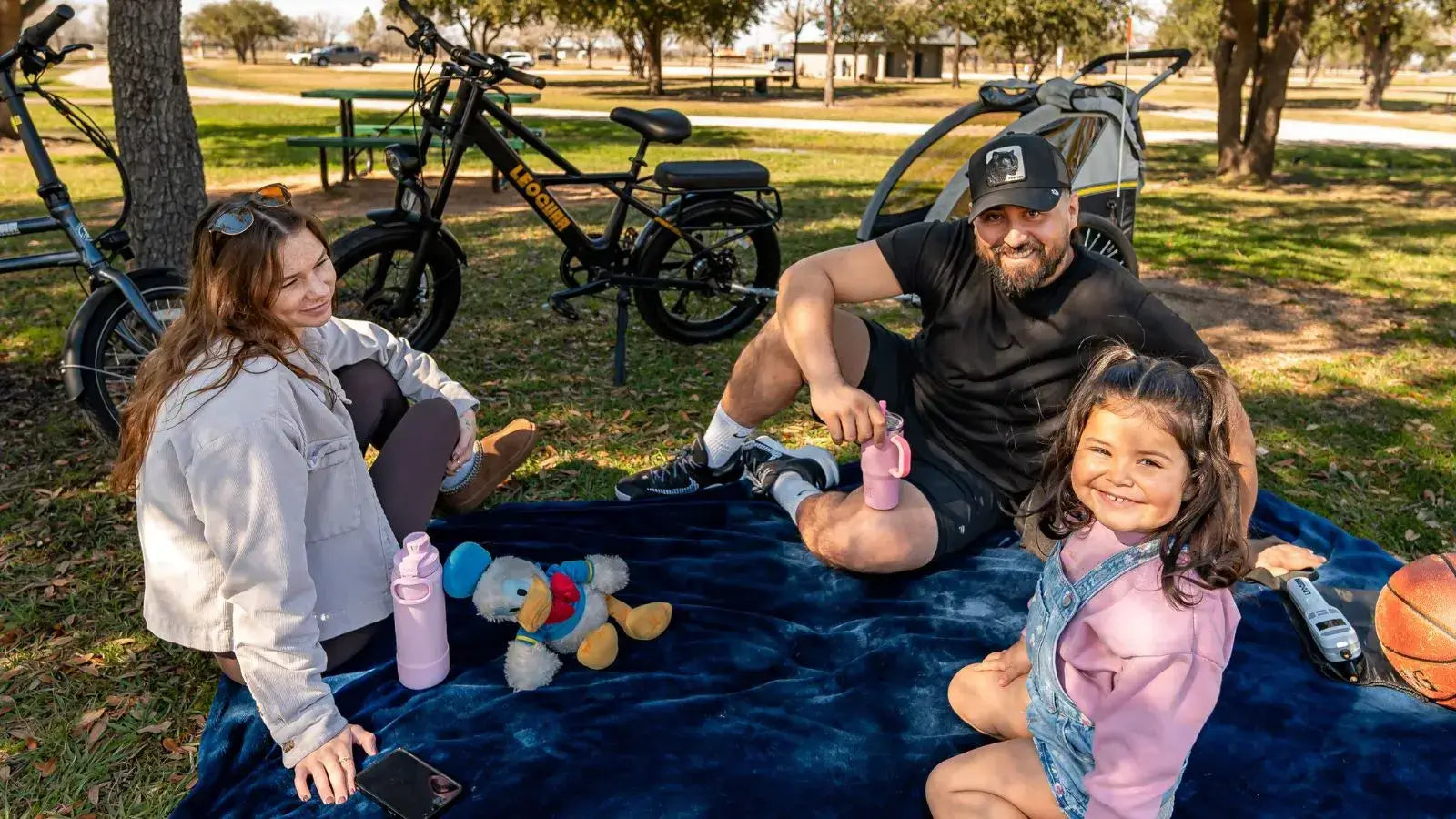 Parents riding an electric cargo bike with a child in a trailer near a playground