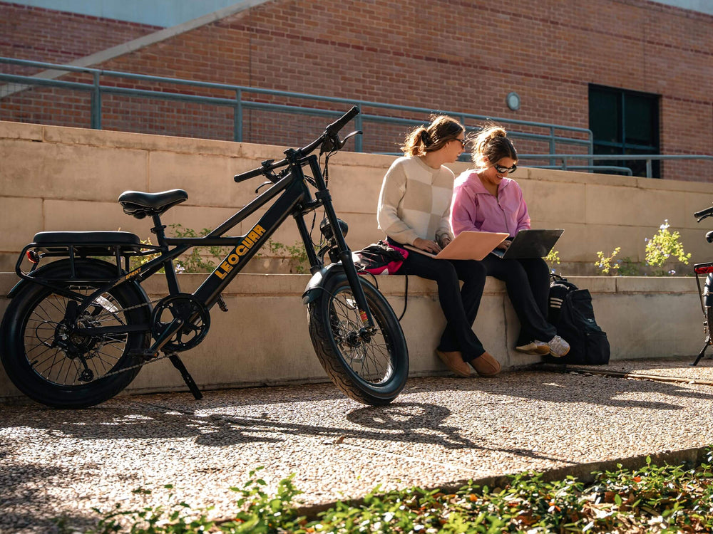 Two women using a laptop and a tablet next to a Sprint Fat Tire Utility Ebike on a sunny day.
