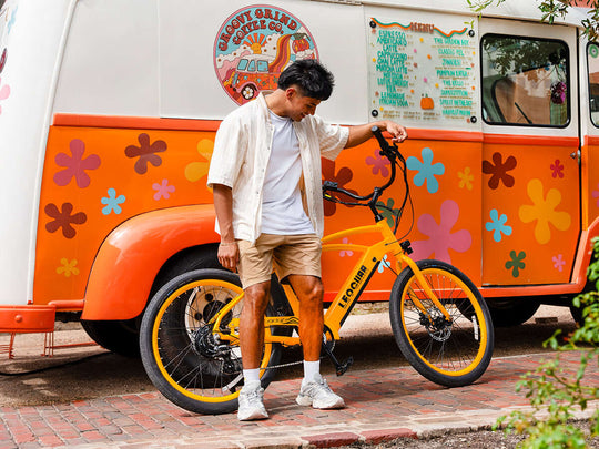 Man standing beside yellow Zephyr SO beach cruiser electric bike in front of a retro orange coffee van