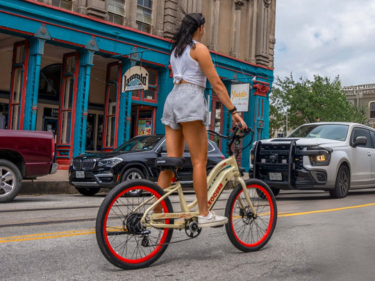 A woman riding a cream Zephyr ST e-bike on a city street with colorful buildings and cars in the background.