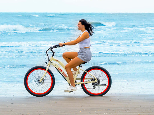 A woman riding a cream-colored Zephyr ST electric cruiser bike along the shoreline with ocean waves in the background.